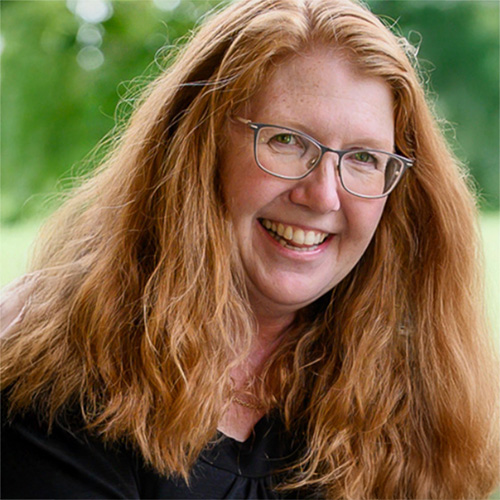 Portrait of Deena Vinger, the owner and lead designer of Inkwell Printers, smiling outdoors with long red hair and glasses.