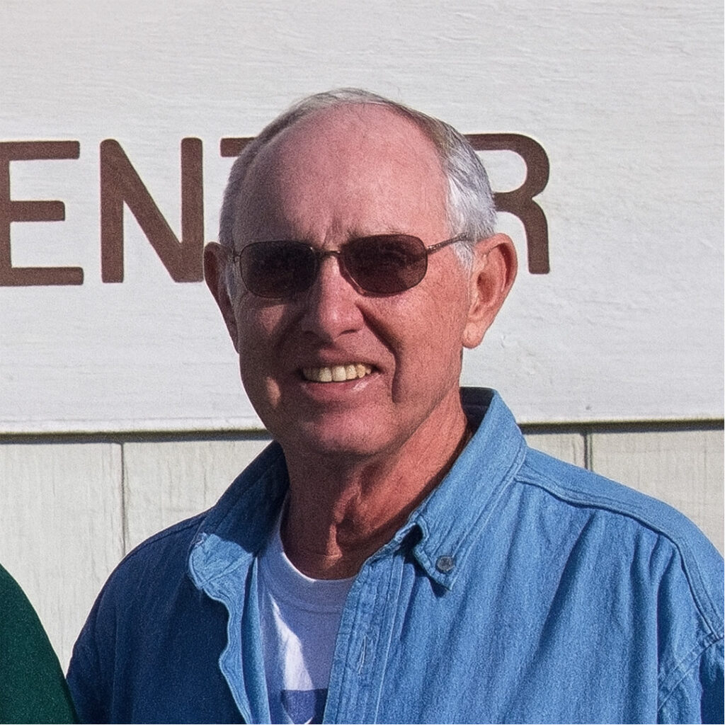 Portrait of an older man wearing sunglasses and a denim shirt, smiling in front of a white wall.