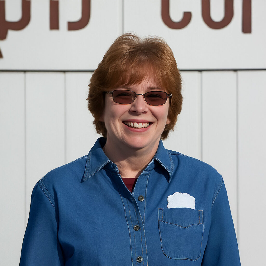 Portrait of a smiling woman wearing sunglasses and a denim shirt with the Inkwell Printers logo, standing in front of a white wall.