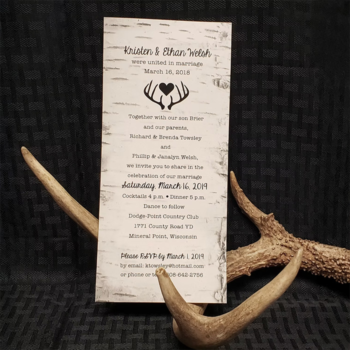 Rustic wedding invitation displayed upright against a dark textured background, with antlers arranged around the card.